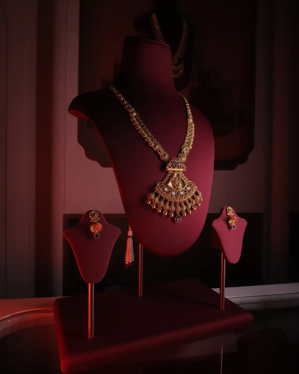 Long temple necklace with a Lakshmi pendant edged in pearls and red drops, with matching earrings, on a red velvet bust