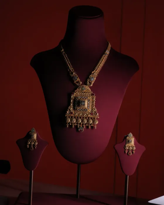 Gold necklace with a large square filigree pendant set with green and red stones, with square drop earrings, on a maroon velvet bust