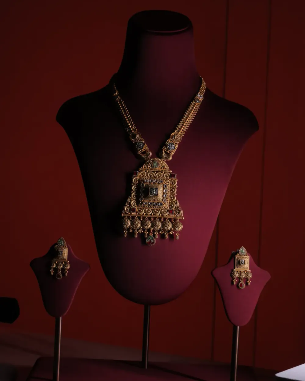Gold necklace with a large square filigree pendant set with green and red stones, with square drop earrings, on a maroon velvet bust